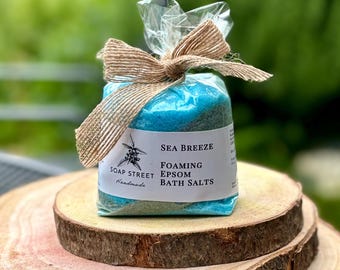 May include: A blue and green bath salt mixture in a clear plastic bag tied with a brown ribbon. The bag has a white label that reads "Sea Breeze Foaming Epsom Bath Salts" and "Soap Street Handmade".