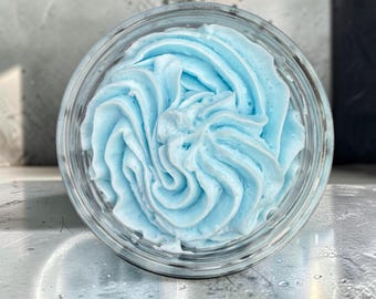 May include: A clear glass jar filled with light blue whipped cream or body butter. The substance is piped in a swirling, decorative pattern that rises above the rim of the container, showcasing a smooth, airy texture with distinct ridges.