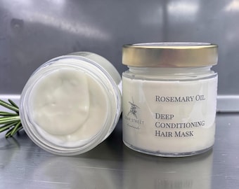 May include: A clear glass jar filled with white cream, labeled Rosemary Oil Deep Conditioning Hair Mask by Soap Street, featuring a gold lid and a sprig of green rosemary next to an open jar showing the thick, smooth white hair mask texture.