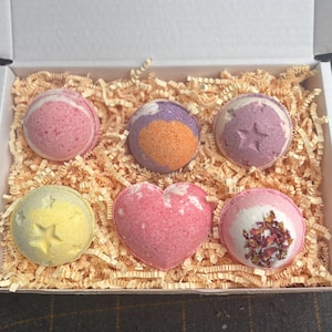 May include: A box filled with six colorful bath bombs. The bath bombs are round, heart-shaped, and decorated with stars and dried rose petals. The box is filled with shredded paper for cushioning.