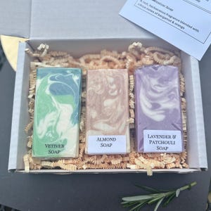 May include: A gift box containing three rectangular soaps with marbled designs. The soaps are labeled "Vetiver Soap", "Almond Soap", and "Lavender & Patchouli Soap". The box is filled with shredded paper for cushioning.