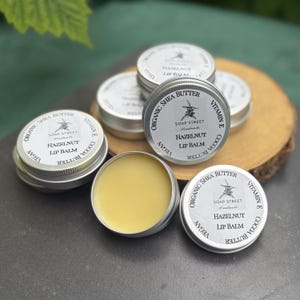 May include: A collection of small, silver lip balm tins with white labels, some stacked, on a wooden surface. One tin is open, showing a light yellow balm. Labels read "Organic Shea Butter, Hazelnut Lip Balm, Vitamin E, Cocoa Butter" and "Soap Street."