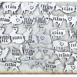 May include: A collection of silver metal charms with various animal shapes and vegan-related text, including "Vegan Chick", "Not a Nugget", "Cruelty Free", "Vegan", "Custom", "Ahimsa", and "Friend Food".