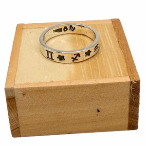 May include: A silver band ring featuring engraved black zodiac symbols including Gemini, Sagittarius, and Aquarius, along with star shapes. The interior of the band is stamped with the text STERLING and the letters OR.