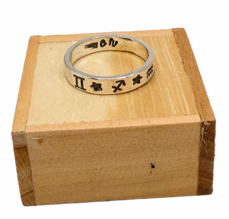 May include: A silver band ring featuring engraved black zodiac symbols including Gemini, Sagittarius, and Aquarius, along with star shapes. The interior of the band is stamped with the text STERLING and the letters OR.