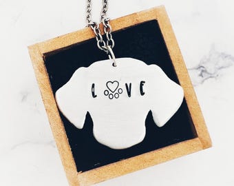 May include: A silver-colored dog-shaped pendant necklace with the word "I LOVE" stamped on it. The pendant is on a silver chain and set against a black background within a wooden frame.