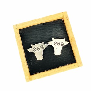 Op de afbeelding: Twee zilveren koe-vormige labels, elk met het nummer "269" in een vetgedrukte letter, worden weergegeven op een zwart oppervlak in een houten frame. De labels zijn naast elkaar geplaatst en het frame heeft een lichtbruine kleur.