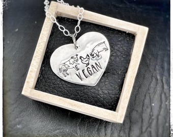 May include: A silver heart-shaped pendant necklace with a cow, chicken, and pig design. The pendant also has the word "VEGAN" engraved on it.