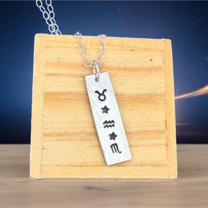 May include: A silver-colored pendant necklace featuring a rectangular tag with stamped zodiac symbols: Taurus, star, Aquarius, star, and Scorpio. The pendant hangs from a delicate silver chain. The necklace is displayed on a light brown wooden block.