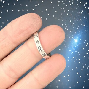 May include: A silver ring featuring engraved zodiac symbols and stars. The ring is shown on a finger against a starry background. The ring's design includes the Taurus, star, Aquarius, and other zodiac signs.