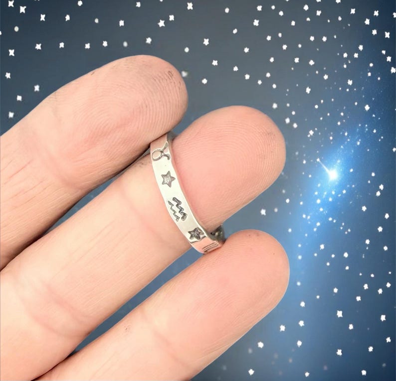 May include: A silver ring featuring engraved zodiac symbols and stars. The ring is shown on a finger against a starry background. The ring's design includes the Taurus, star, Aquarius, and other zodiac signs.