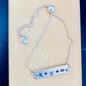 May include: Silver chain bracelet with a silver bar charm featuring the zodiac signs for Scorpio, Taurus, and Aquarius. The bracelet has a sliding bead closure.