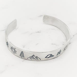 May include: A silver-colored cuff bracelet with engraved outdoor-themed designs. The bracelet features a sun, pine tree, mountain range, tent, and other nature-inspired illustrations. The open-ended design allows for easy wear and removal.