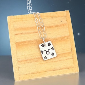 May include: Silver-toned pendant necklace featuring a rectangular charm with a Taurus zodiac symbol and star accents. The charm hangs from a delicate chain. The background is a wooden surface against a blue backdrop with stars.