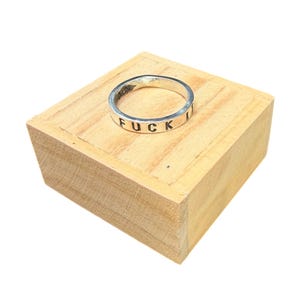 May include: A silver ring with the word "FUCK IT" engraved around the band. The ring is displayed on a small, light-colored wooden block, set against a white background. The ring's design is simple and bold.