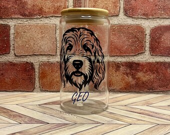 May include: A clear glass tumbler with a bamboo lid and a curved glass straw. The tumbler has a black dog face graphic and the name "GEO". The background is a brick wall and a wooden surface.