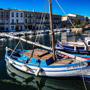 May include: A small sailboat, painted blue and white, is moored in a canal. The boat features a wooden deck and mast, with the registration number ST 374 852 visible. Other boats and buildings are visible along the canal.