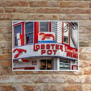 May include: A red and white sign with the words "Lobster Pot" in red neon letters. The sign is attached to a building with red and white siding. There is a lobster illustration on the sign.