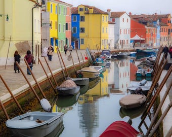 Può includere: Una vista pittoresca di un canale fiancheggiato da edifici colorati a Burano, Italia. Piccole barche sono ormeggiate lungo il canale, con pali di legno. Le persone camminano sul marciapiede e l'acqua riflette gli edifici.