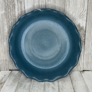 May include: A round, teal-colored plate with a textured surface and a scalloped edge. The plate is made of ceramic and is suitable for serving food. The plate is a solid color and has a matte finish.