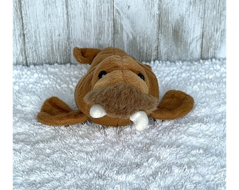 May include: A brown plush walrus toy with white tusks. The walrus is lying on a white fluffy surface.