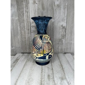 May include: A tall, decorative vase with a flared top and a rounded body. The vase features a dark blue textured top and a cream-colored body with a hand-painted design of a bird and fish in black, yellow, and red. The vase is approximately 10 inches tall.