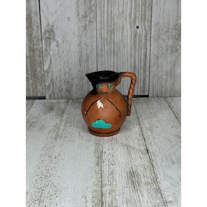 May include: A small, brown ceramic pitcher with a black rim and handle. The pitcher is decorated with turquoise cloud and feather designs. The handle has a rope-like texture.