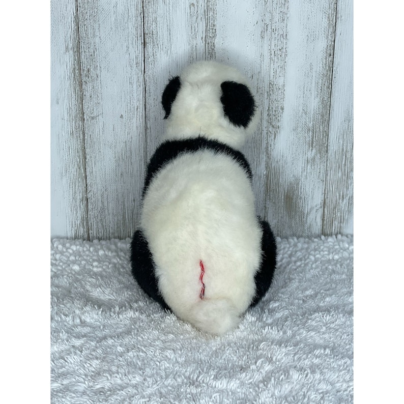 May include: A black and white plush panda bear sitting on a white fluffy surface. The panda has a black nose and black eyes.