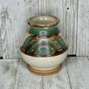 May include: A small, handcrafted ceramic vase with a wide base and a narrow opening. The vase features a cream-colored base, with a band of green and brown geometric designs around the middle. The rim and base are a darker brown color.