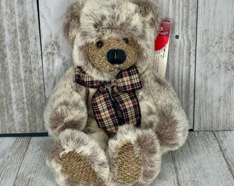 May include: A plush teddy bear with a light brown and gray fur texture. The bear has a plaid bow tie with red and brown accents and a brown nose. The bear is sitting on a wooden surface.