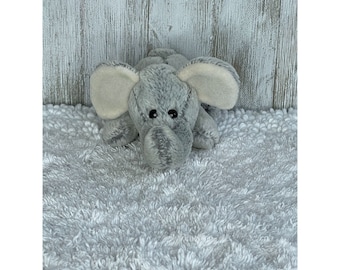 May include: A gray plush elephant toy with large ears and black eyes. The elephant is lying on a white fluffy surface.