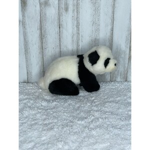 May include: A black and white plush panda bear toy sitting on a white fluffy surface. The panda is lying on its side with its head turned to the right.