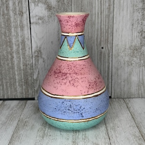 May include: A decorative ceramic vase with a bulbous shape and a flared top. The vase features a color scheme of pink, blue, and green, accented with gold trim. The design includes geometric patterns and speckled textures, creating a vintage aesthetic.