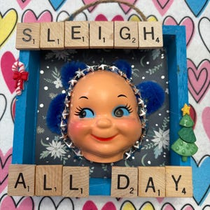 Puede incluir: Una obra de arte enmarcada con una cara de muñeca vintage con orejeras azules y una diadema con estrellas. Las palabras "SLEIGH ALL DAY" están escritas con letras de madera. Un bastón de caramelo y un árbol de Navidad adornan el marco azul.