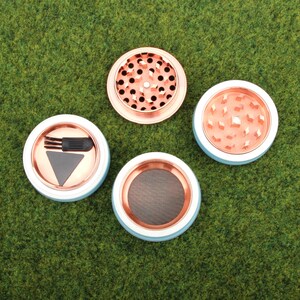 May include: Four parts of a metal grinder with a blue and white design. The grinder has a screen, a compartment for herbs, a compartment for kief, and a scraper.