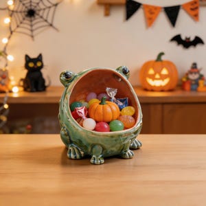 Puede incluir: Un cuenco de cerámica verde con forma de rana y la boca muy abierta, lleno de caramelos de colores variados y una pequeña figura de calabaza naranja. La rana tiene un acabado brillante, ojos visibles en la parte superior y pies esculpidos.