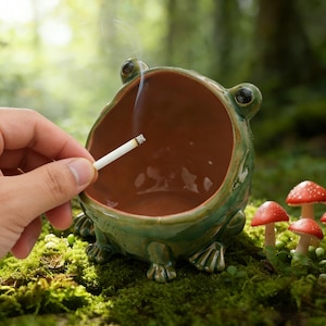 Pode incluir: Um cinzeiro de cerâmica verde em forma de sapo com interior castanho. O cinzeiro tem um design caprichoso com patas de sapo e olhos grandes. Um cigarro aceso é segurado acima do cinzeiro. Figuras de cogumelos vermelhos no fundo.