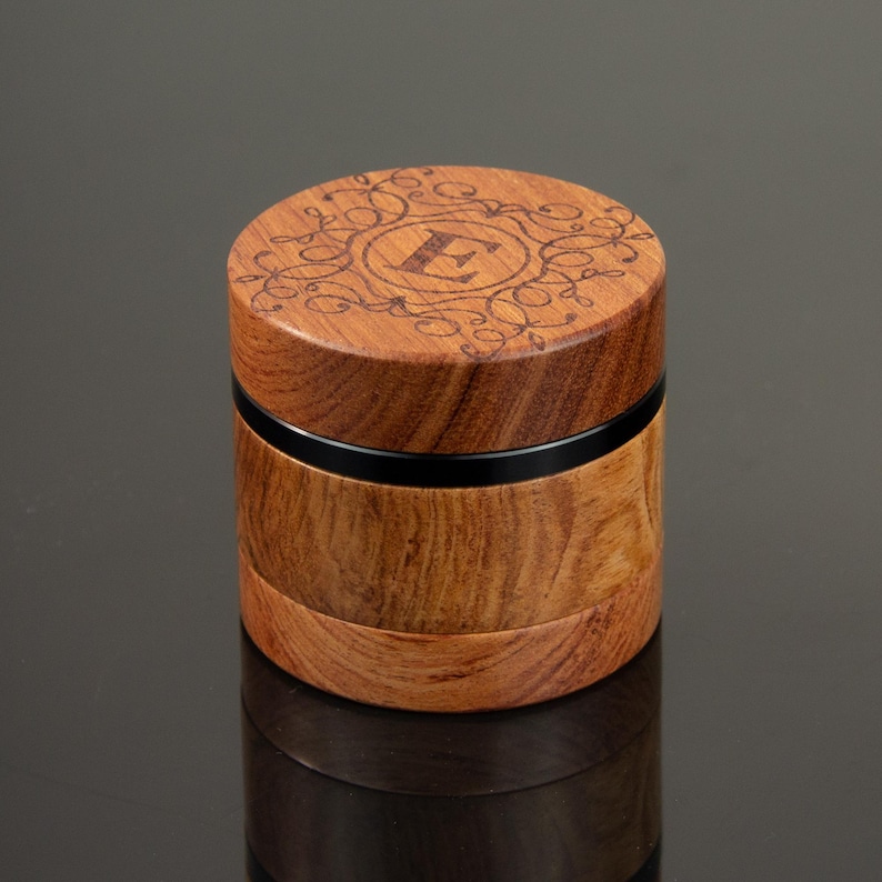 May include: A cylindrical wooden grinder with a natural brown wood grain finish. The top lid features a laser-engraved letter E surrounded by an intricate filigree pattern. A thin, smooth black band separates the top and bottom sections of the device.