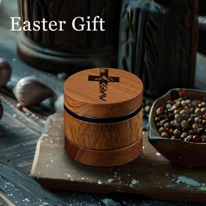 May include: A cylindrical wooden grinder with a dark brown wood grain finish. The top lid features a laser-engraved design of a cross, a mountain range, and the text "GOD > G > AV". The text "Easter Gift" is visible in the upper left corner.