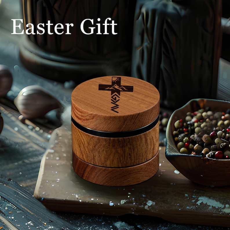 May include: A cylindrical wooden grinder with a dark brown wood grain finish. The top lid features a laser-engraved design of a cross, a mountain range, and the text "GOD > G > AV". The text "Easter Gift" is visible in the upper left corner.