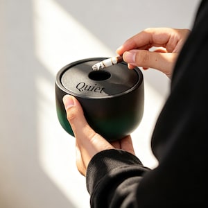 May include: A black and green ashtray with the word "Quiet" printed on the side. The ashtray is round with a hole in the center. A lit cigarette is being placed in the ashtray.