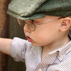 Puede incluir: Un niño pequeño con una gorra de tweed verde y gafas redondas con montura dorada. El niño también lleva una camisa abotonada a rayas. El niño está tocando una superficie de madera.