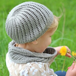 以下が含まれることがあります： グレーのニット帽とそれに合わせたマフラーをつけた小さな子供が、黄色のタンポポの花を持っている