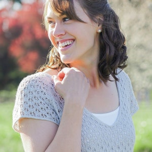 Puede incluir: Una mujer con cabello castaño y ojos azules sonríe mientras lleva una blusa de crochet gris claro y pantalones vaqueros azules.