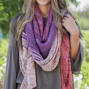 May include: A woman wearing a gray long-sleeved shirt with a purple, pink, and orange shawl. She is standing in front of a green tree and a blurred background.