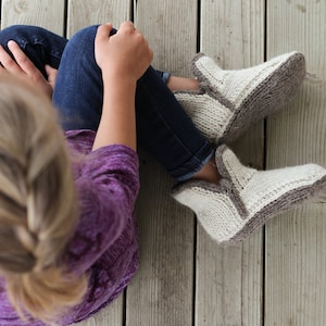 May include: A person wearing a purple sweater and blue jeans sits on a wooden floor wearing a pair of knitted gray and white slippers.