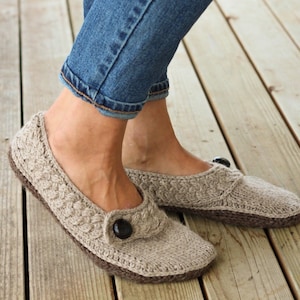 Pode incluir: Um par de pantufas de malha bege com um detalhe de botão. As pantufas têm um design trançado e são usadas em um piso de madeira.