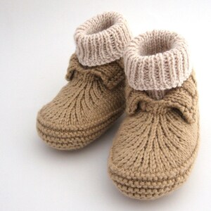 May include: A pair of brown knitted baby booties with a beige knitted cuff. The booties have a bow detail on the front.