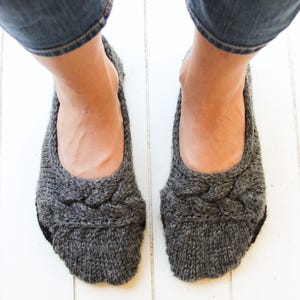 May include: A pair of gray knitted slipper socks with a braided design. The socks are worn on a person's feet.