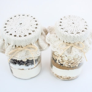 May include: Two glass jars filled with baking ingredients, topped with crocheted lids and tied with twine bows. The jars are on a white surface.